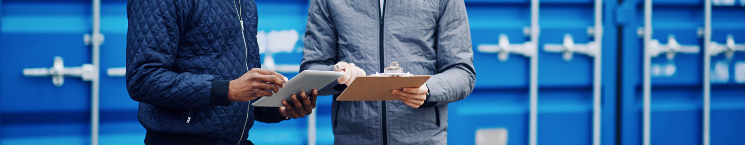 Two men holding tablets in front of blue shipping containers