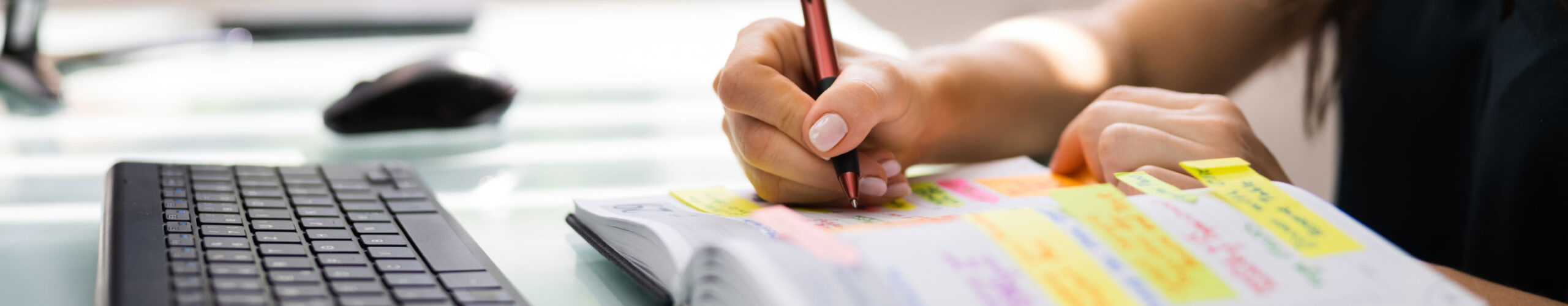 Close up of woman writing in agenda with sticky notes in front of computer keyboard