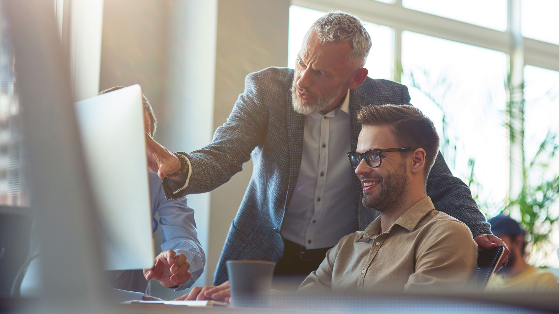 Older man leaning over the shoulder of a younger male employee reviewing a computer monitor