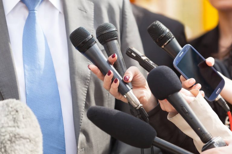 reporters holding microphones to businessman at press conference